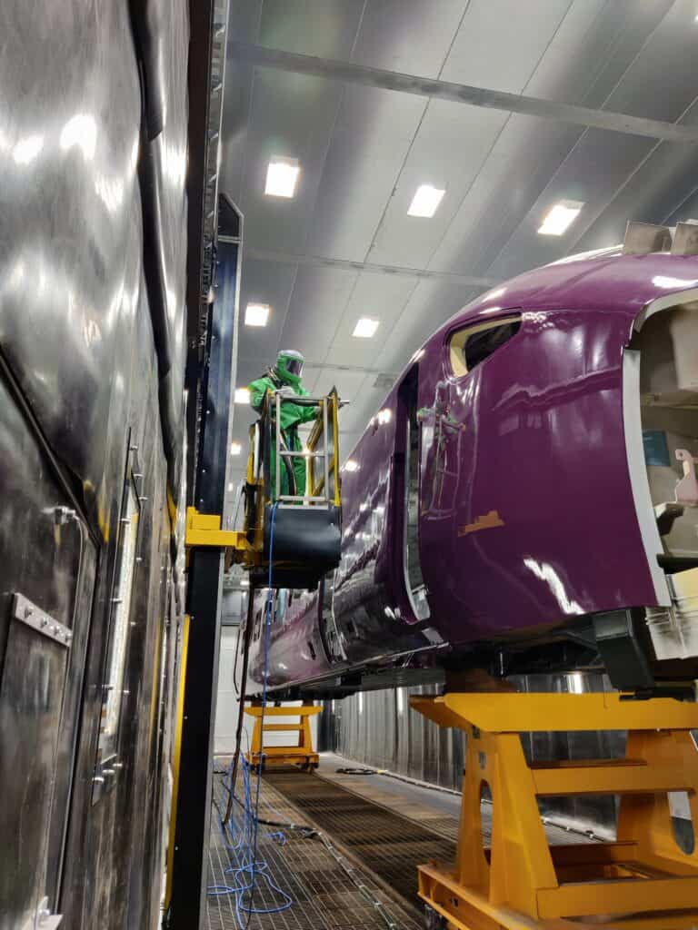 Blast operator in full GVS RPE blasting PPE holding a blast hose and nozzle processing rolling stock inside the blast room at Hitachi Rail.
